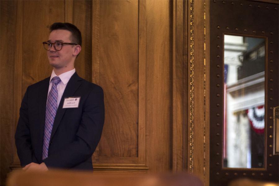 A student wears a suit and tie and stands against a wood paneled wall.